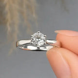 A silver solitaire engagement ring with a round-cut clear gemstone, held between two fingers against a soft gray background with blurred beige dried grass.